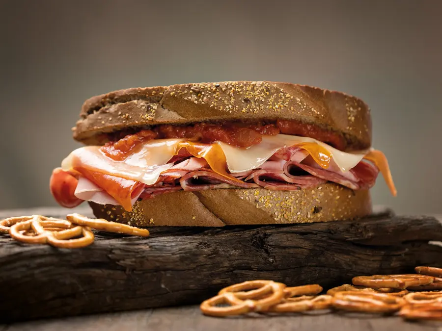 A toasted deli sandwich stacked with ham and cheese on a dark board with pretzels