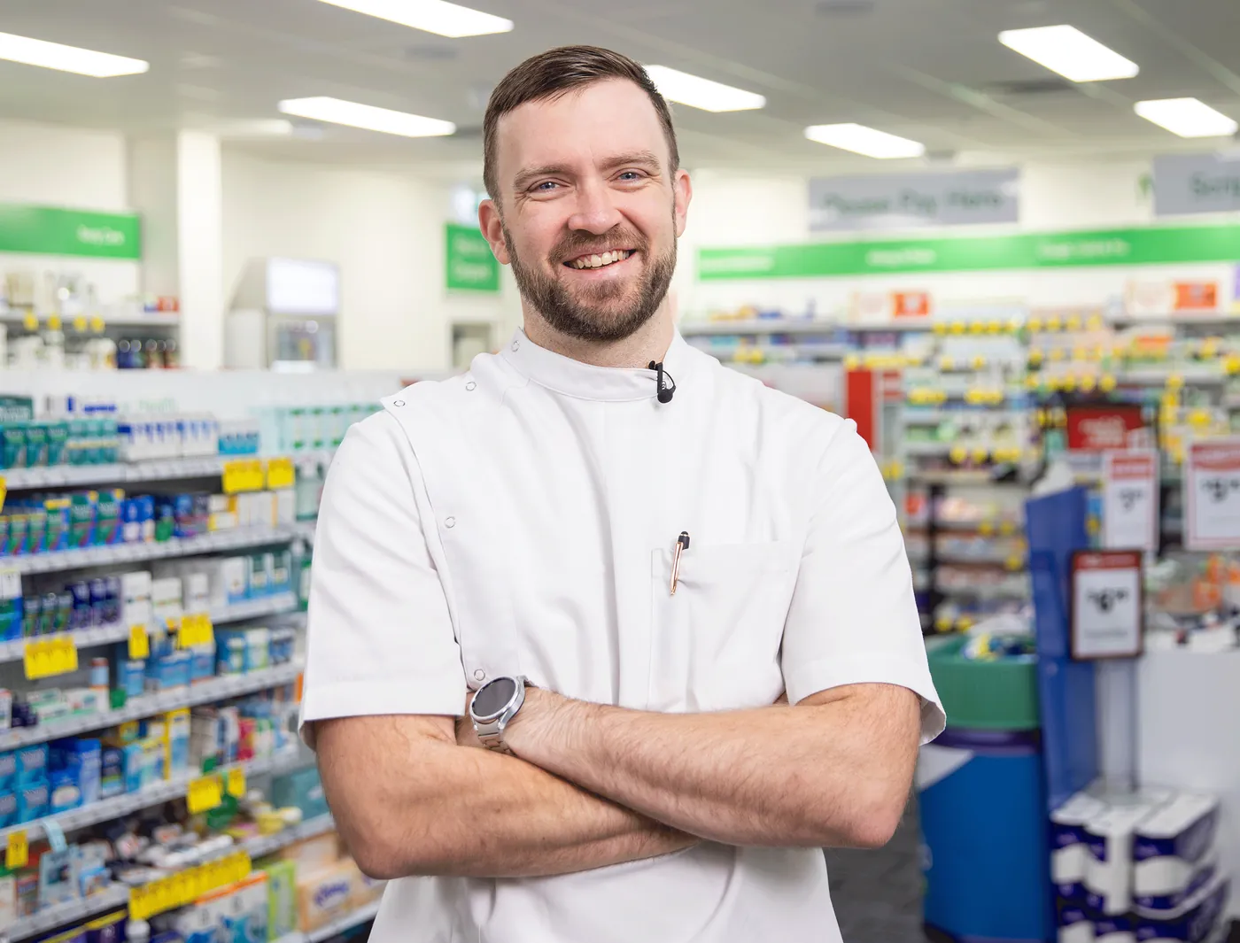 A smiling pharmacist with arms crossed in the aisle of his pharmacy