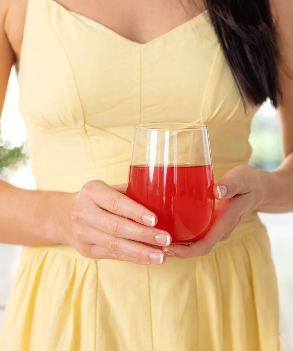 Close up of hands holding a glass of raspberry collagen drink against a yellow summer dress