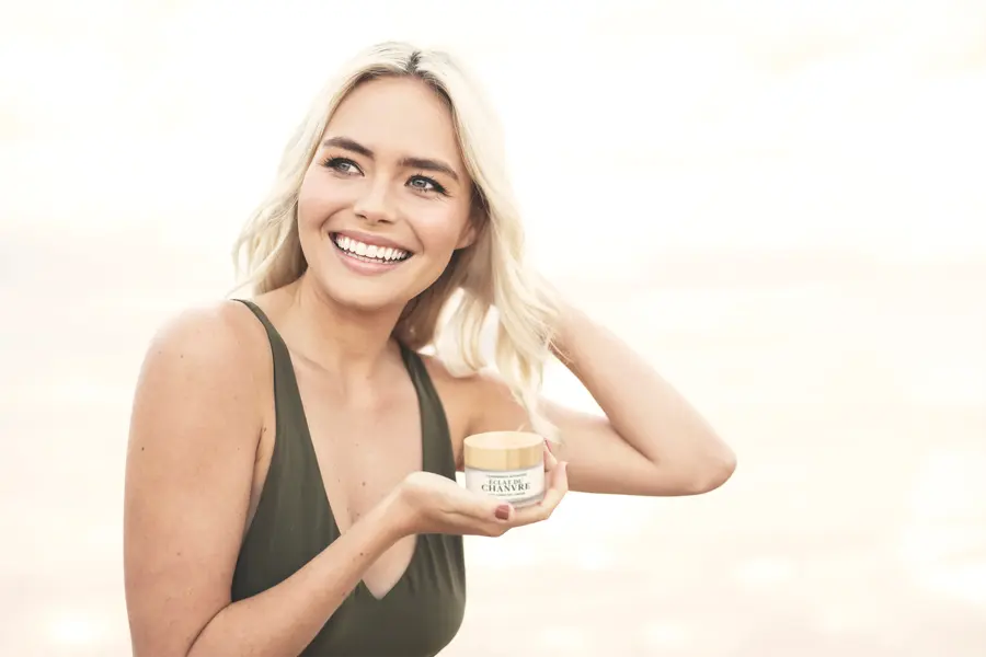 A smiling woman in a green swimsuit holding a small jar of cream at the beach
