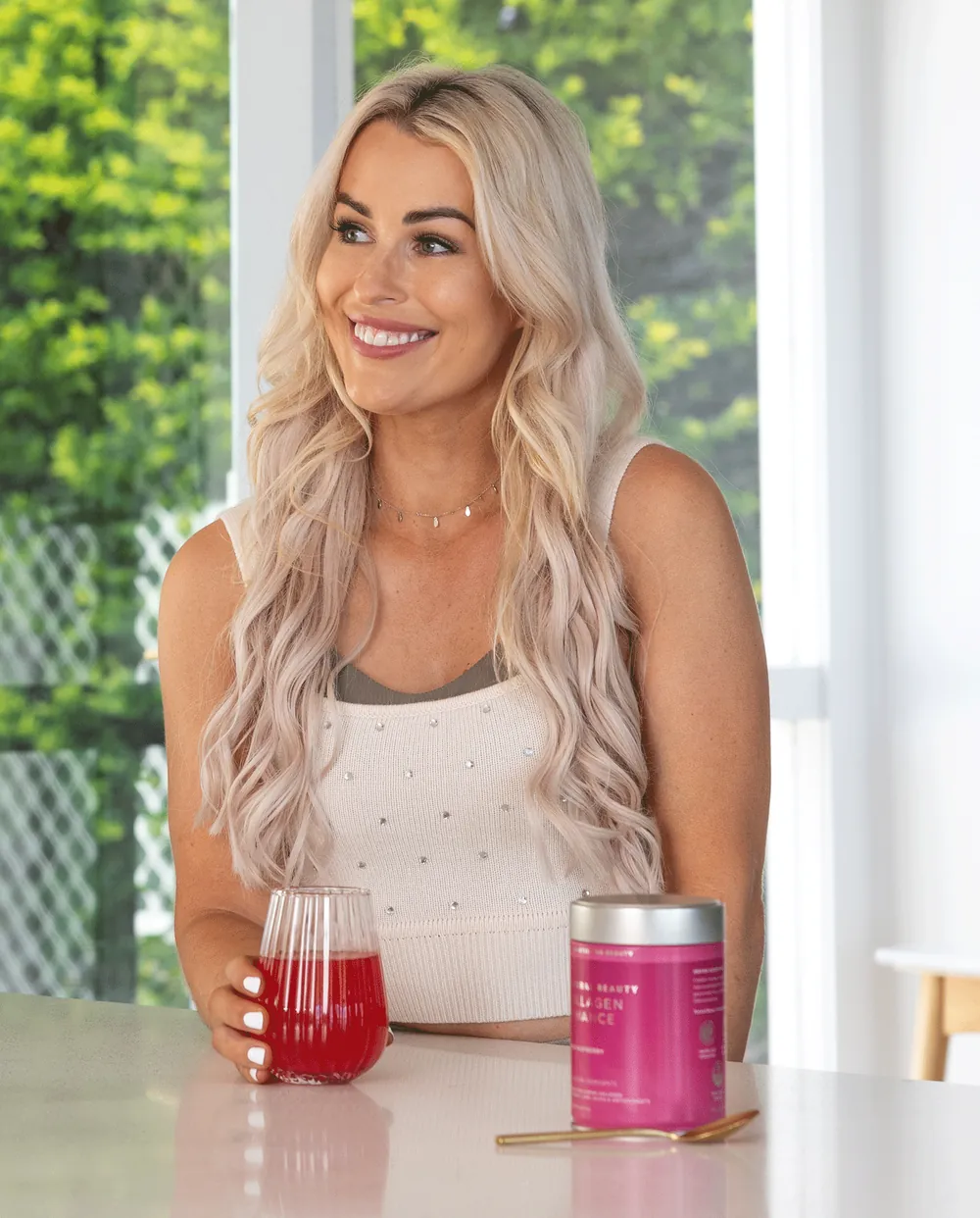 A woman at a kitchen bench with a glass of raspberry collagen drink beside a Collagen Enhance tin