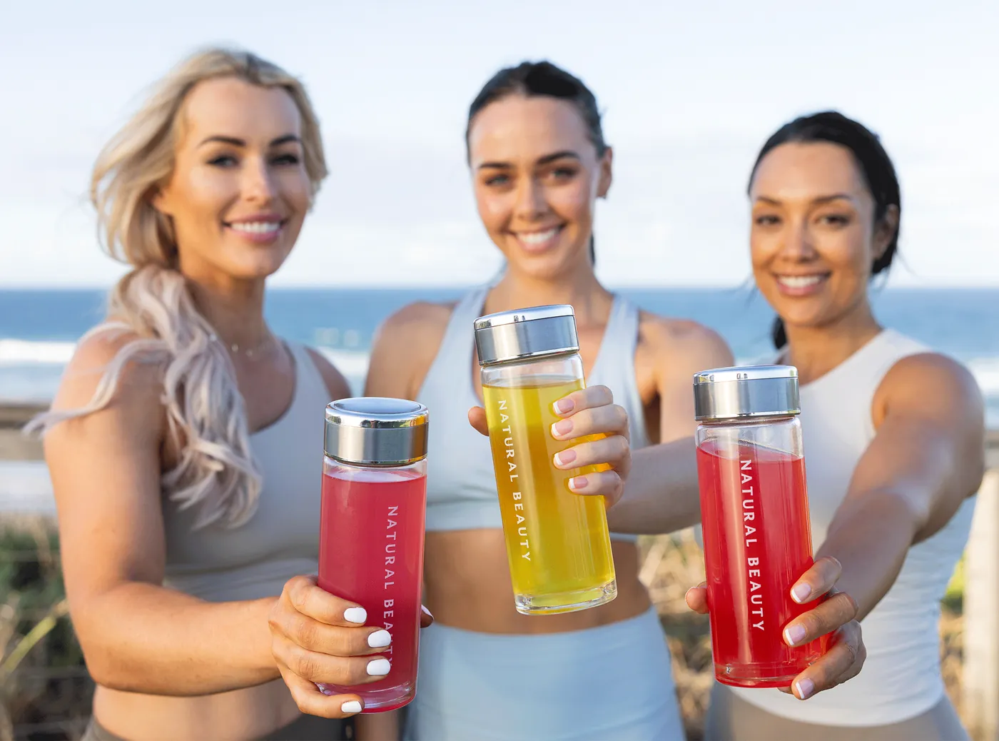 Three women by the ocean holding Natural Beauty bottles out towards the camera