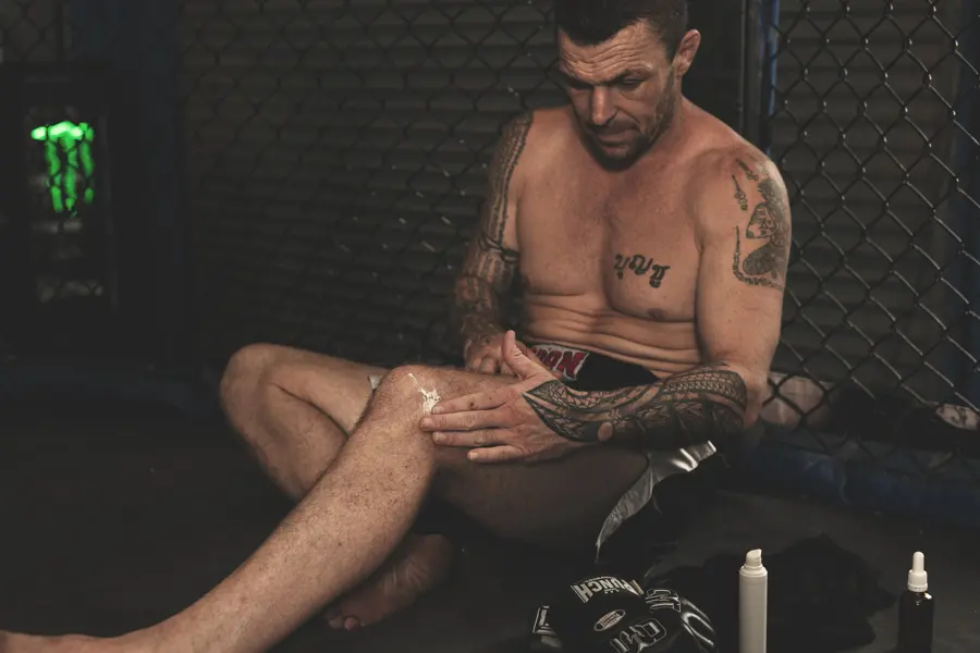 A fighter sitting against the cage applying cream to his leg